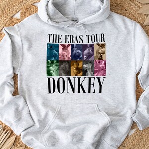 Pode incluir: Camisola de moletom cinza com capuz com um gr&aacute;fico de Donkey de Shrek. O gr&aacute;fico apresenta uma colagem do rosto de Donkey em diferentes express&otilde;es. O texto "The Eras Tour" e "Donkey" est&atilde;o impressos em preto.