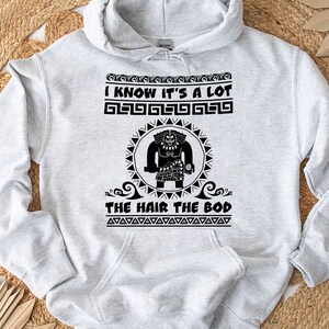 May include: Gray hooded sweatshirt with a black graphic print of a cartoon character with the text "I know it's a lot the hair the bod" printed on the front.