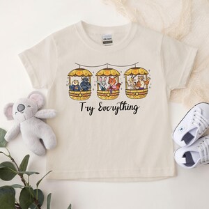 Pode incluir: Camiseta creme com um gr&aacute;fico colorido de tr&ecirc;s g&ocirc;ndolas, cada uma com personagens de animais. O texto "Try Everything" est&aacute; impresso abaixo. Um coala de pel&uacute;cia e t&ecirc;nis brancos tamb&eacute;m est&atilde;o na imagem.