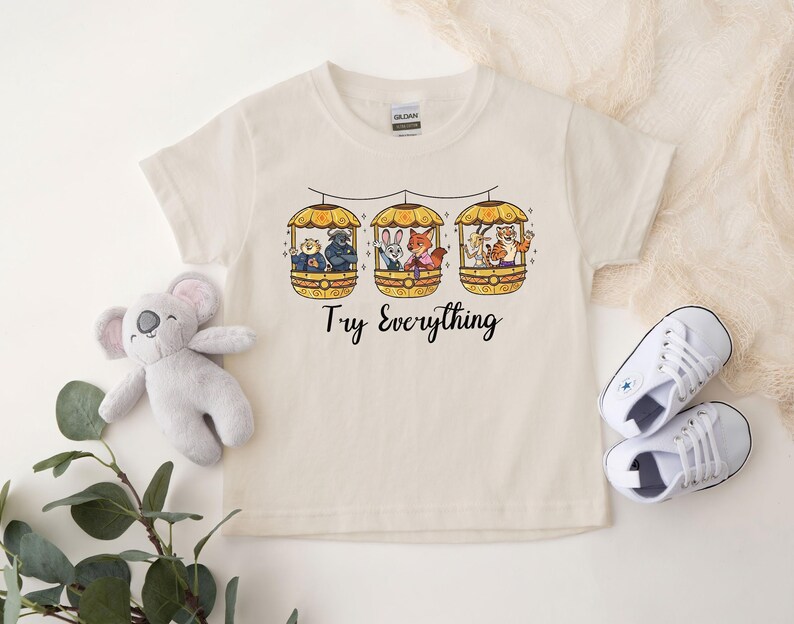Pode incluir: Camiseta creme com um gr&aacute;fico colorido de tr&ecirc;s g&ocirc;ndolas, cada uma com personagens de animais. O texto "Try Everything" est&aacute; impresso abaixo. Um coala de pel&uacute;cia e t&ecirc;nis brancos tamb&eacute;m est&atilde;o na imagem.