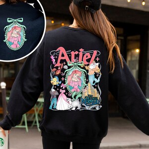 Pu&ograve; includere: Felpa nera a maniche lunghe con una stampa grafica di Ariel de La Sirenetta. La grafica presenta Ariel, Flounder, Sebastian e re Tritone. Il testo "Ariel" e "La Sirenetta" &egrave; incluso anche nella grafica.