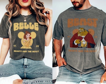 Può includere: Una coppia di t-shirt in stile vintage con personaggi del film Disney La Bella e la Bestia. La maglietta a sinistra presenta un'illustrazione gialla e marrone di Belle con il testo "Belle" e "La Bella e la Bestia". La maglietta a destra presenta un'illustrazione marrone della Bestia con il testo "Beast".