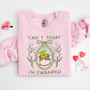 Puede incluir: Sudadera rosa con un personaje de ogro verde en un marco con el texto "Can't today I'm swamped".