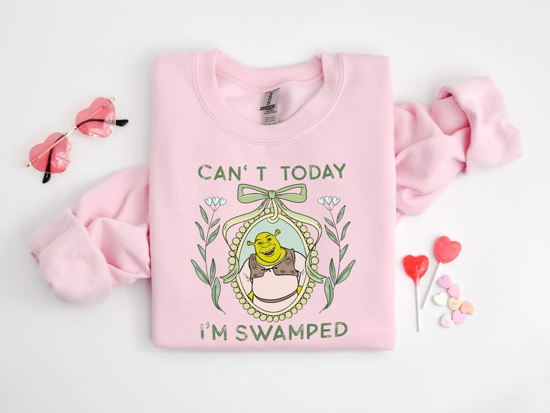 Puede incluir: Sudadera rosa con un personaje de ogro verde en un marco con el texto "Can't today I'm swamped".