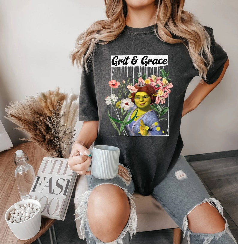 May include: A dark grey t-shirt featuring a graphic print of a green ogre character with flowers and the text "Grit & Grace".