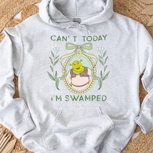 Puede incluir: Una sudadera con capucha gris con el texto "Can't today I'm swamped" y una imagen de dibujos animados de Shrek en un marco con flores.