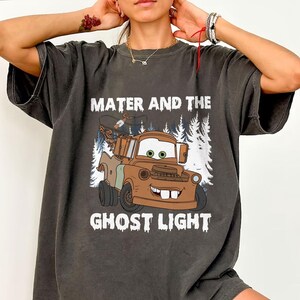 May include: Dark grey t-shirt with a cartoon image of Mater from the film Cars, and the text "Mater and the Ghost Light". The graphic depicts Mater, a brown tow truck with a friendly face, set against a backdrop of white trees.