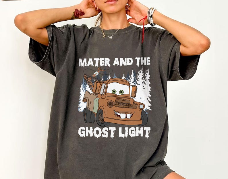 May include: Dark grey t-shirt with a cartoon image of Mater from the film Cars, and the text "Mater and the Ghost Light". The graphic depicts Mater, a brown tow truck with a friendly face, set against a backdrop of white trees.