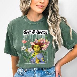 May include: A green t-shirt featuring a graphic of a green ogre wearing a crown, surrounded by flowers. The text "Grit & Grace" is printed above the graphic.