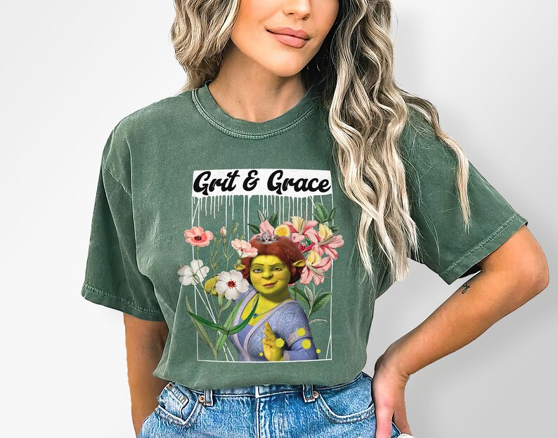 May include: A green t-shirt featuring a graphic of a green ogre wearing a crown, surrounded by flowers. The text "Grit & Grace" is printed above the graphic.
