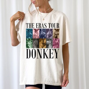 Pode incluir: Uma camiseta branca com uma estampa gr&aacute;fica de um burro do filme Shrek. O gr&aacute;fico &eacute; uma colagem de diferentes imagens do burro em v&aacute;rias cores. O texto "THE ERAS TOUR DONKEY" &eacute; impresso abaixo do gr&aacute;fico.
