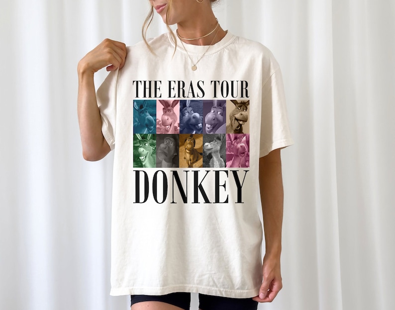 Pode incluir: Uma camiseta branca com uma estampa gr&aacute;fica de um burro do filme Shrek. O gr&aacute;fico &eacute; uma colagem de diferentes imagens do burro em v&aacute;rias cores. O texto "THE ERAS TOUR DONKEY" &eacute; impresso abaixo do gr&aacute;fico.