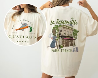 May include: A cream-coloured t-shirt featuring a graphic of Remy the rat from the film Ratatouille. The front of the shirt reads "Anyone can cook" with a rat holding a carrot. The back of the shirt reads "La Ratatouille" with a graphic of the restaurant from the film. The text "Paris, France" is below the graphic.