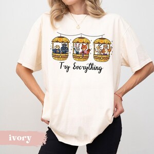 Pode incluir: Camiseta cor marfim com tr&ecirc;s g&ocirc;ndolas amarelas com personagens de desenhos animados. Abaixo das g&ocirc;ndolas, o texto "Try Everything" est&aacute; escrito em preto. A camiseta &eacute; usada por uma pessoa.