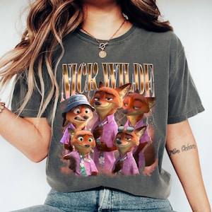 May include: Dark gray t-shirt featuring a graphic of Nick Wilde from Zootopia. The design includes multiple cartoon foxes in pink shirts, with the text "NICK WILDE" in gold lettering. The shirt is a comfortable, casual style.