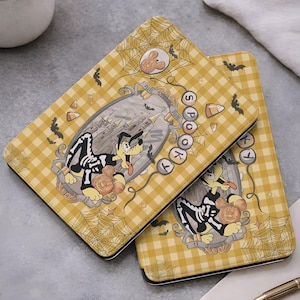 May include: A yellow and white gingham patterned e-reader case featuring a cartoon dog wearing a skeleton costume, a castle illustration, spiderwebs, bats, candy corn, and the text "SPOOKY" arranged vertically in circular bubbles.