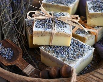 Puede incluir: Barras de jabón de lavanda hechas a mano con una base amarilla clara y una capa superior de capullos de lavanda secos. Cada barra está atada con cinta de rafia. Una cuchara de madera con lavanda y ramitas de lavanda secas también están en la imagen.