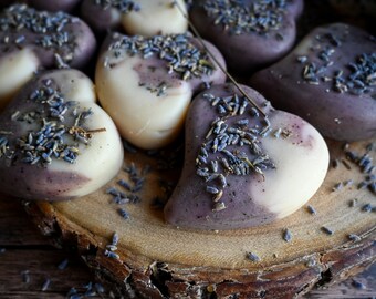 May include: Heart-shaped soaps with a two-tone design, lavender and cream, are arranged on a wooden surface. The soaps are topped with dried lavender buds. The soaps are a natural color and are likely handmade.