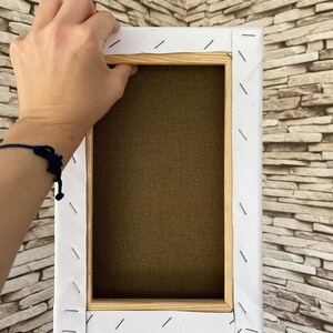 May include: The back of a stretched canvas with a wooden frame. The canvas is white and has been stapled to the frame. The frame is made of natural wood.