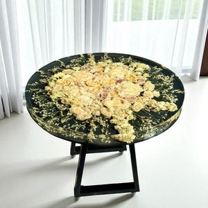 May include: A round table with a black base and a top embedded with dried flowers. The flowers are primarily cream and yellow, arranged in a floral pattern. The table is approximately 30 inches in diameter.