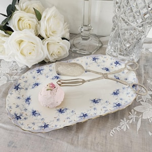 May include: An oval serving platter with a blue and white floral pattern and gold edging. Silver serving tongs and a small pink dessert with a rose are on the platter. White roses and a crystal vase are in the background.