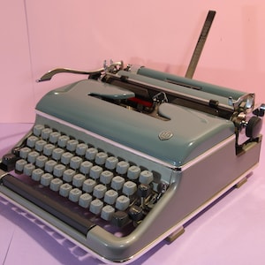 Op de afbeelding: Een vintage blauwe typemachine met een zilveren retourwagenhendel en een zwart metalen frame. De typemachine heeft een standaard toetsenbordindeling met ronde toetsen.