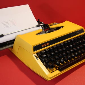 May include: A bright yellow Brother Deluxe 220TR portable typewriter with a black keyboard, a red key, and a white sheet of paper inserted into the carriage. The device features a black control panel with the brand name printed in orange text.
