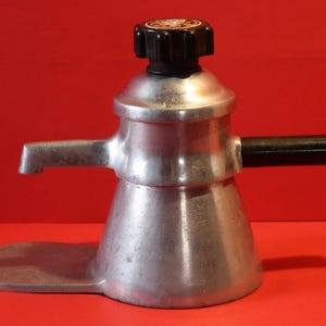 Könnte beinhalten: Eine silberne Kaffeemaschine aus Aluminium mit einem schwarzen Griff und einem schwarzen Knopf oben. Die Kaffeemaschine hat einen Ausguss an der Seite und eine Basis, die breiter ist als die Oberseite.
