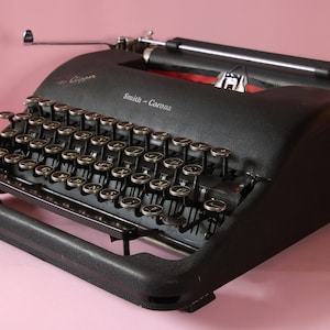 May include: A black Smith-Corona Clipper typewriter with silver keys and accents. The vintage machine has a textured surface and the brand name is visible on the front. The typewriter sits on a pink background.