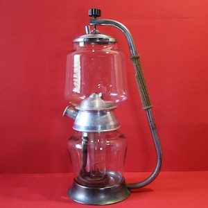 May include: A vintage glass and metal soda siphon with a long, curved metal tube. The siphon has a clear glass body and a silver-colored metal base. The tube is attached to the top of the siphon and has a black knob.