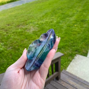 May include: A multi-colored fluorite crystal carving in shades of blue, green, and purple. The polished stone has a smooth, curved shape, held in a hand against a backdrop of green grass and a wooden deck.