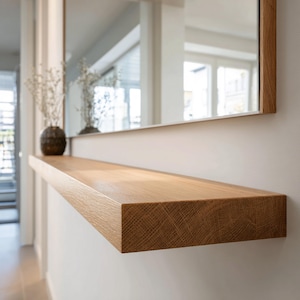 May include: A rectangular, light-brown wooden shelf mounted on a white wall, with a mirror above. The shelf is made of solid wood and has a natural grain pattern. A decorative vase with dried flowers sits on the shelf.