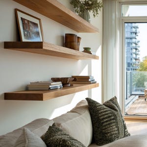 May include: Three wooden floating shelves mounted on a white wall, displaying a framed picture, books, and decorative items. A sofa with pillows is in the foreground, and a window with a view of a building is in the background.