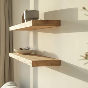 May include: Two floating wooden shelves mounted on a white wall. The shelves are made of light-colored wood and have a natural grain. The top shelf holds a gray textured vase and a small wooden object. The bottom shelf holds a white bowl with decorative items.
