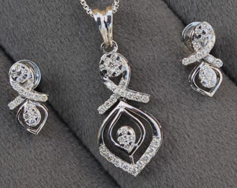 May include: A silver necklace and earrings set with a delicate pendant featuring a diamond-shaped design with a smaller diamond in the center. The pendant is surrounded by smaller diamonds.