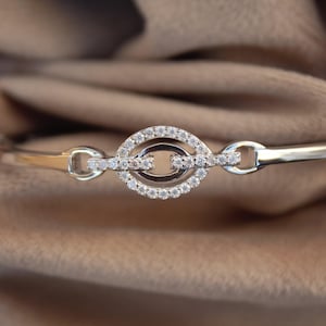 Puede incluir: Un brazalete de plata con un diseño de cristal transparente. El brazalete presenta un diseño de doble círculo con un círculo más pequeño dentro del círculo más grande. El círculo más pequeño tiene una línea recta en el centro.