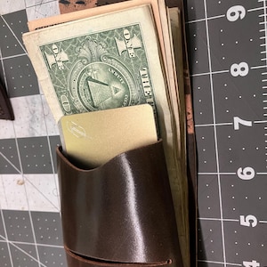 May include: A dark brown leather wallet containing several US one-dollar bills and a gold-colored metal card with a silver logo. The wallet is positioned on a gray cutting mat with white grid lines and visible inch markings along the right side.