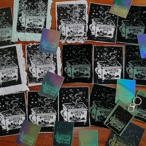 May include: A collection of black and white hand-carved woodblock prints featuring a steaming bowl of soup with the text "Dumpster Soup". The prints are on a wooden surface with some iridescent stickers.