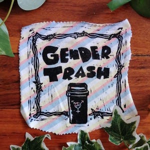 May include: A fabric patch with the words "GENDER TRASH" in bold black letters, framed by a barbed wire design. The patch features a trash can with a gender symbol, set against a striped background of blue, pink, and yellow.
