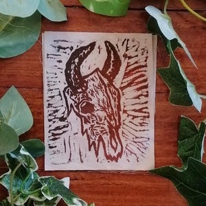 Könnte beinhalten: Ein braun-weißer Linolschnitt eines gehörnten Schädels. Das Kunstwerk zeigt eine detaillierte Darstellung eines Schädels mit großen Hörnern vor einem strukturierten Hintergrund. Der Druck befindet sich auf einem quadratischen Papierstück, umgeben von grünen Blättern.