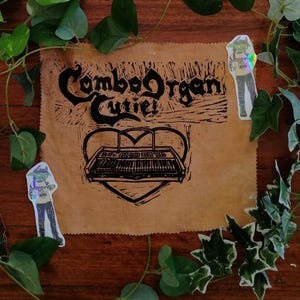 May include: A brown fabric featuring the text "Combo Organ Cutie!" and a black and white keyboard illustration within a heart shape. Two stickers of a cartoon character are placed on the fabric.