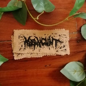 May include: Two rectangular, tan-coloured patches with a jagged edge, stacked on top of each other. The patches have black text that is difficult to read. The patches are on a wooden surface with green leaves.