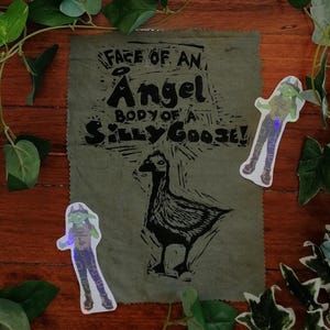 May include: A distressed green fabric with the text "FACE OF AN ANGEL BODY OF A SILLY GOOSE!" and a black and white goose illustration. Two holographic stickers of a goblin are placed on the fabric.