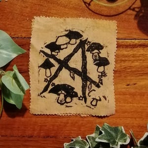 Peut inclure: Impression sur tissu beige carré avec un pentagramme noir orné de champignons et de feuilles. L'impression a un aspect rugueux et texturé, avec un bord décoratif et dentelé. L'œuvre est posée sur un fond en bois.