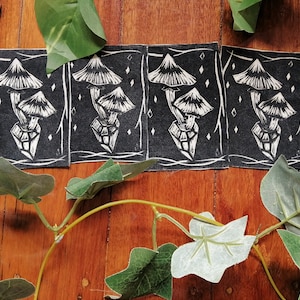 May include: Five black and white linocut prints of mushrooms with a crystal base. The prints are on a wooden surface with green leaves.
