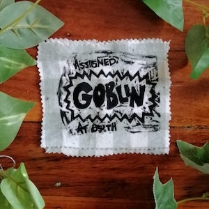 May include: A square, off-white patch with a black graphic that reads "ASSIGNED: GOBLIN AT BIRTH". The patch has a zig-zag border and a checkered pattern. The background is a wooden surface with green leaves.