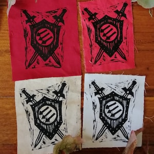 May include: Four fabric patches with a black and white design. Each patch features a shield with three downward arrows, crossed by two swords. Two patches are red, and two are off-white.