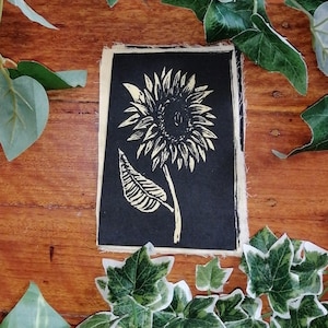 Könnte beinhalten: Ein schwarz-weißes Linolschnitt-Bild einer Sonnenblume. Die Sonnenblume ist auf einem schwarzen Hintergrund zentriert, mit einer detaillierten Darstellung der Blütenblätter, des Stiels und des Blattes der Blume. Der Druck befindet sich auf cremefarbenem Papier mit einem rauen Rand. Der Hintergrund ist eine Holzoberfläche mit grünen Blättern.