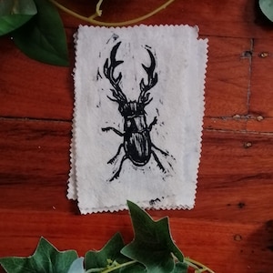 Könnte beinhalten: Ein schwarz-weiß Linolschnitt-Druck eines Hirschkäfers auf einem weißen Stoffquadrat. Der Käfer ist detailliert mit großen Mandibeln und komplizierten Körpersegmenten. Der Stoff hat einen rohen, unfertigen Rand, vor einem hölzernen Hintergrund.