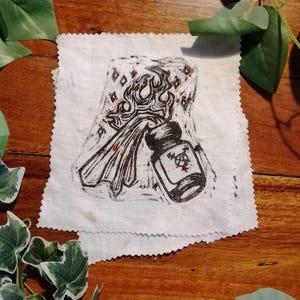 May include: A black and white linocut print on a white fabric square. The print shows a bottle with a symbol and a flame-like design. The fabric has a raw, unfinished edge. The overall theme is mystical or artistic.